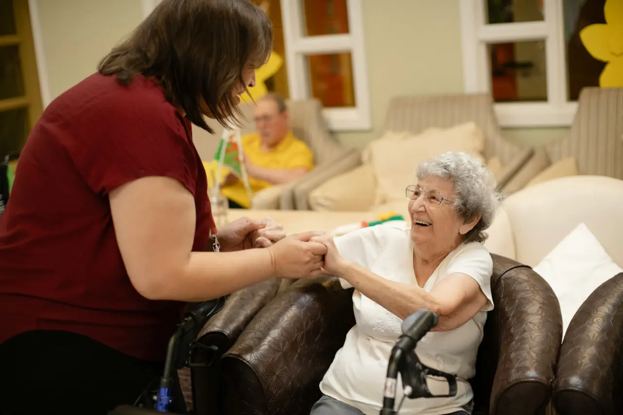 A carer holding hands with a smiling older resident in a care-home lounge